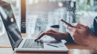 Um homem sentado diante de seu laptop. Com a mão esquerda, ele segura o smartphone, com a direita, usa o laptop. 