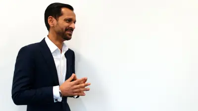 Irfan fica diante da câmera, na frente de uma parede branca. Ele não olha para a câmera. 